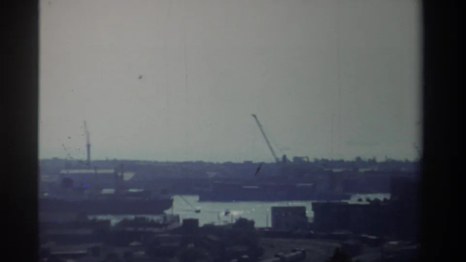 1982 Taiwan home-movie frame showing a distant industrial waterfront with cranes, low structures, and water under a washed, hazy sky.