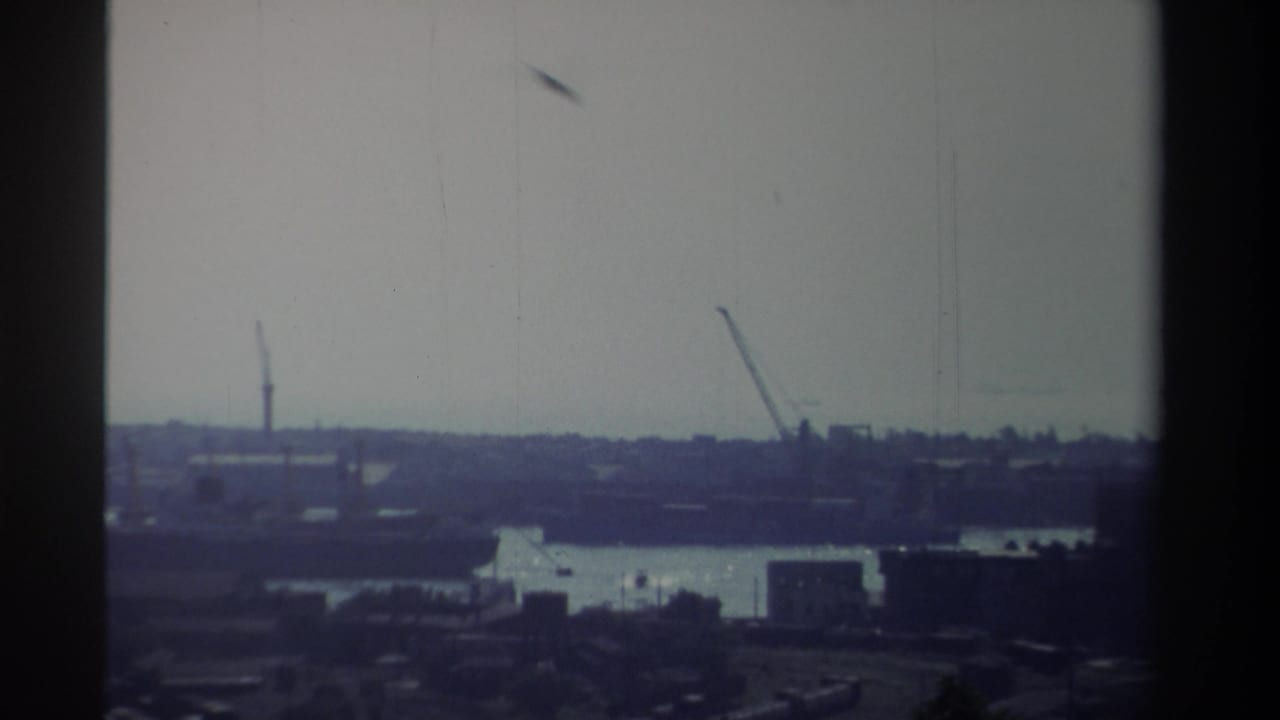 High vantage 8mm frame from Taiwan in 1982 showing distant cranes, a bright waterway, low port buildings, and soft blue-purple film color.