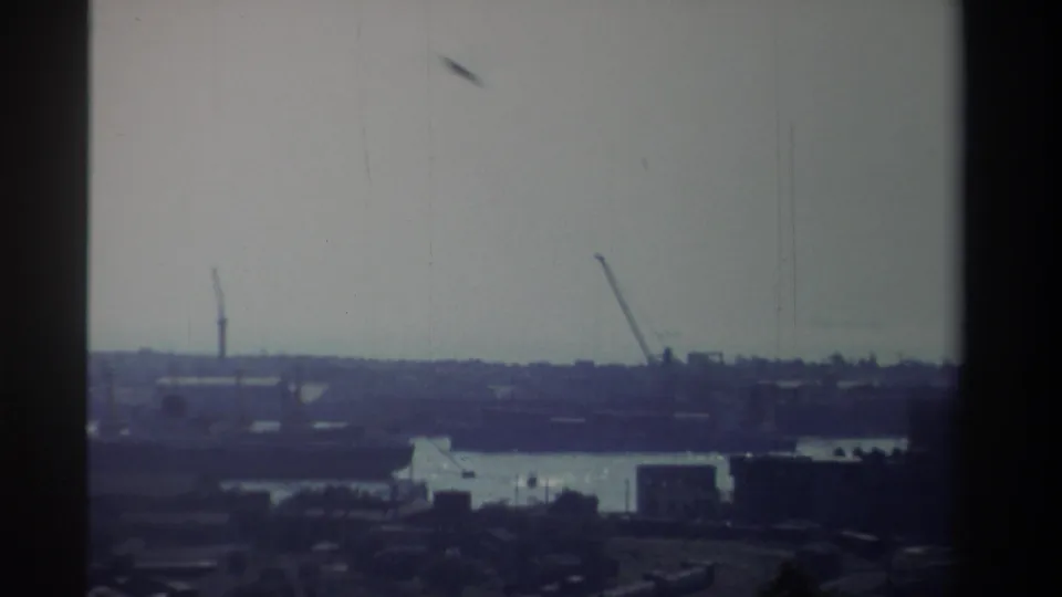 High vantage 8mm frame from Taiwan in 1982 showing distant cranes, a bright waterway, low port buildings, and soft blue-purple film color.