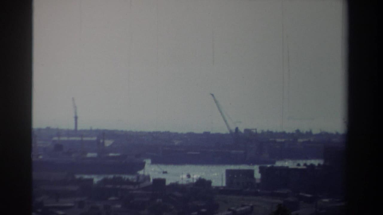 1982 Taiwan home-movie frame showing a soft, hazy industrial waterfront with cranes, water, and distant dock structures.