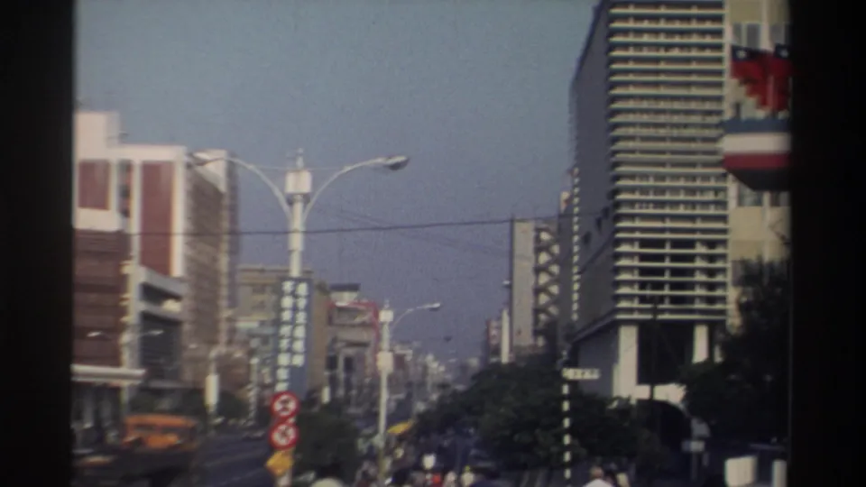 1982 Taiwan street scene with moving cars, modern buildings, streetlights, overhead wires, and a high-rise along the roadway.