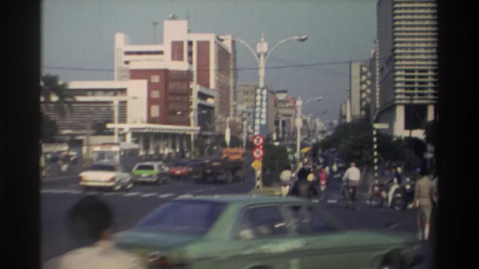 1982 Taiwan city street with blurred foreground car, pedestrians, two-wheel traffic, roadside signs, and modern buildings in soft 8mm footage.