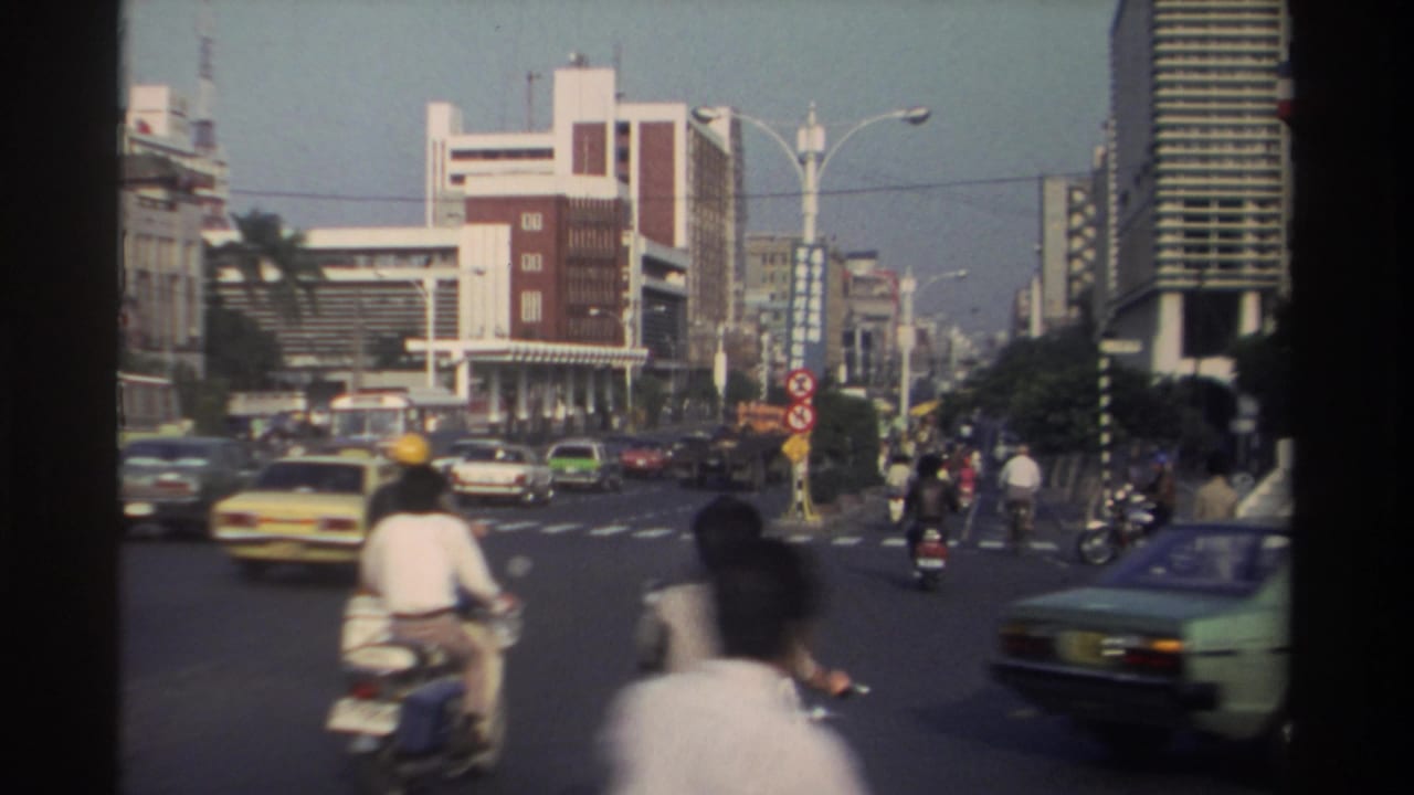 Scooters and cars move through a broad urban intersection with mid-rise buildings ahead.