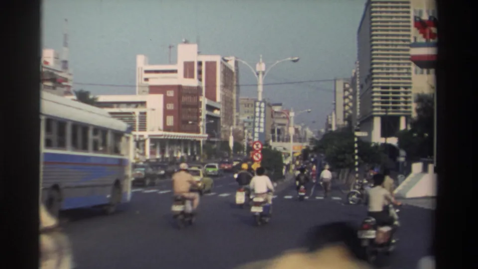 1982 Taiwan urban road view showing a blue-and-white public bus, scooters, cars, pedestrians, and dense mid-rise street architecture.
