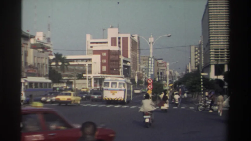 1982 Taiwan city intersection with a bus in the center distance, scooters and cars in motion, pedestrians, signs, and mid-rise buildings.
