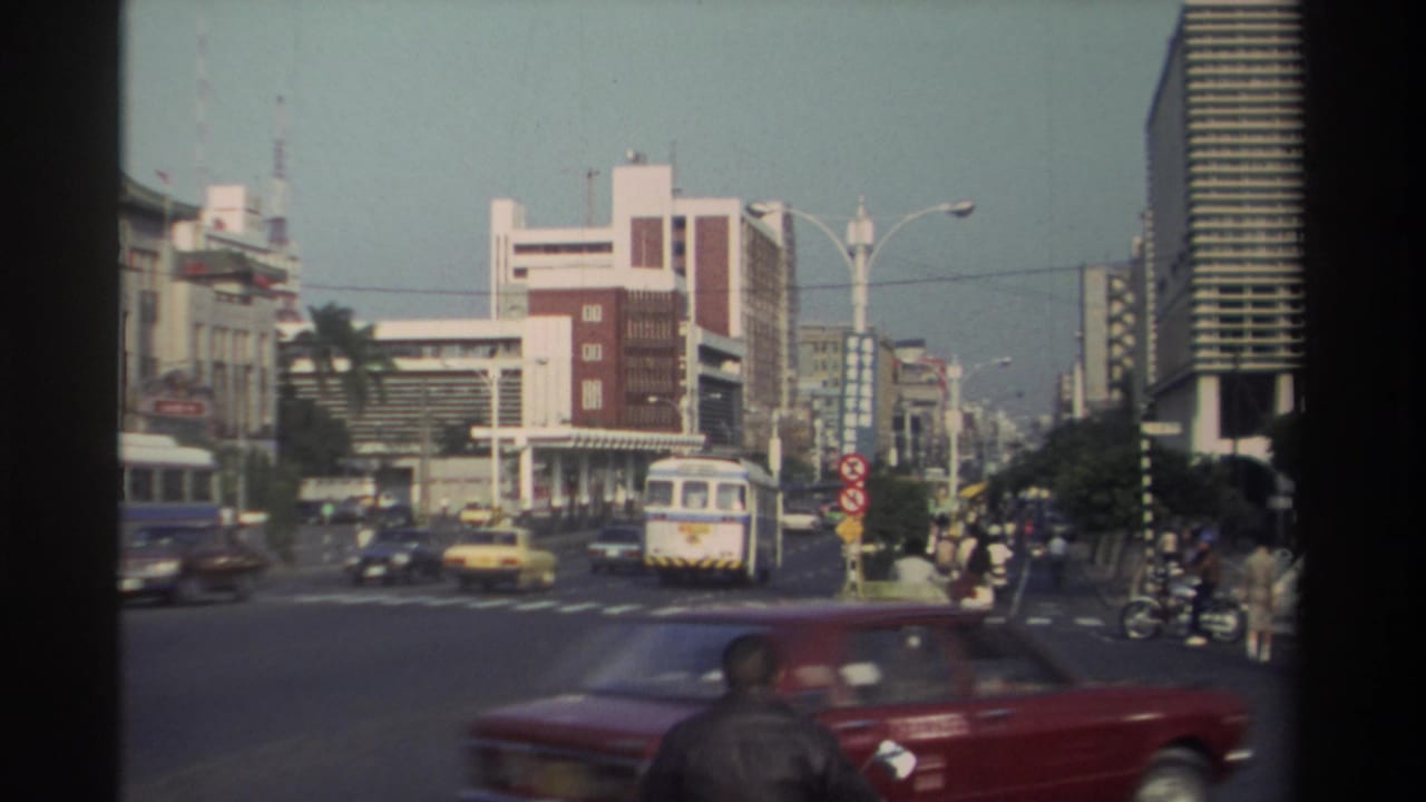 Busy city intersection with cars, a bus, scooters, bicycles, pedestrians, and tall buildings.