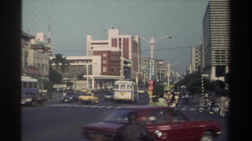 1982 Taiwan street view with a bus, cars, scooters, bicycles, pedestrians, palm trees, and modern buildings at a broad intersection.