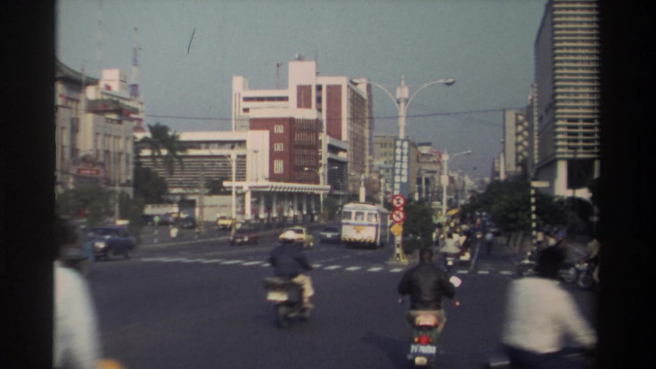 Scooter riders, a white bus, cars, pedestrians, and city buildings at a sunlit intersection.