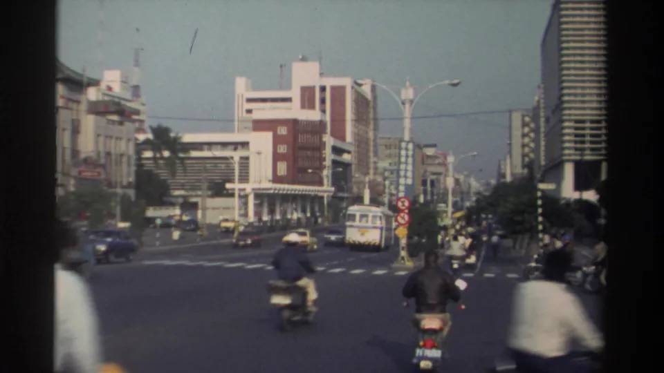 1982 Taiwan intersection with scooter riders in the foreground, a white bus, cars, pedestrians, street signs, and commercial buildings.