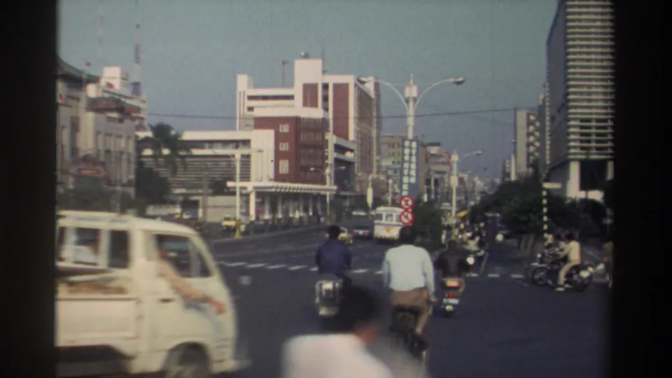 1982 Taiwan street scene with a small light van, bicyclist, scooter riders, pedestrians, cars, palm trees, and buildings around an intersection.