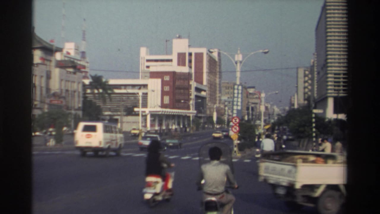 1982 Taiwan street view with scooters and cars crossing a wide intersection, framed by older storefronts and taller modern buildings.
