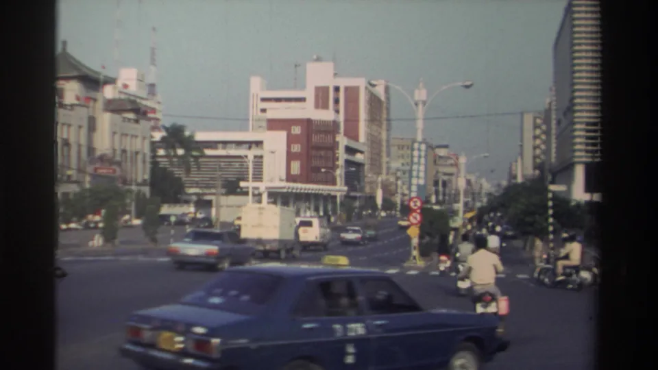 1982 Taiwan city traffic with a blue sedan in the foreground and mixed scooters, vans, and cars crossing a broad road.