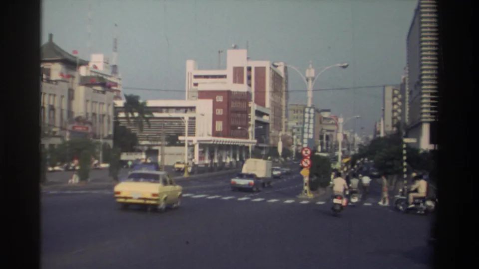 1982 Taiwan intersection view with a yellow car, scooters, pedestrians, and mixed traffic between older and taller buildings.