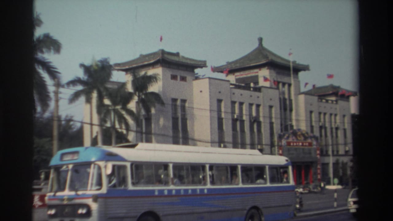 Blue and white bus passing a traditional-style building with flags and palm trees.