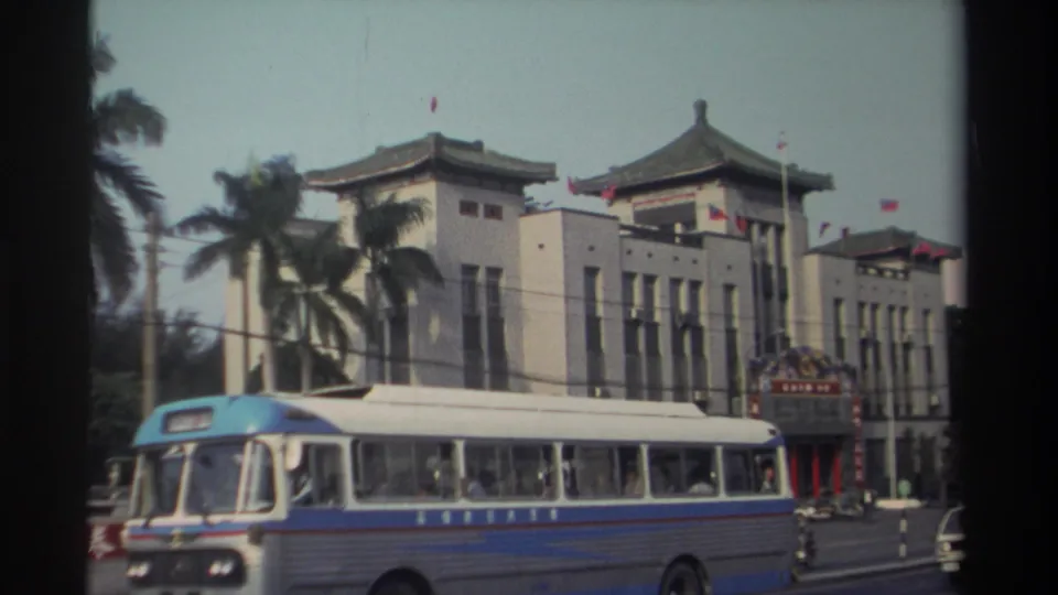 1982 Taiwan street view with a blue and white bus crossing in front of a traditional-style civic building with flags and palm trees.