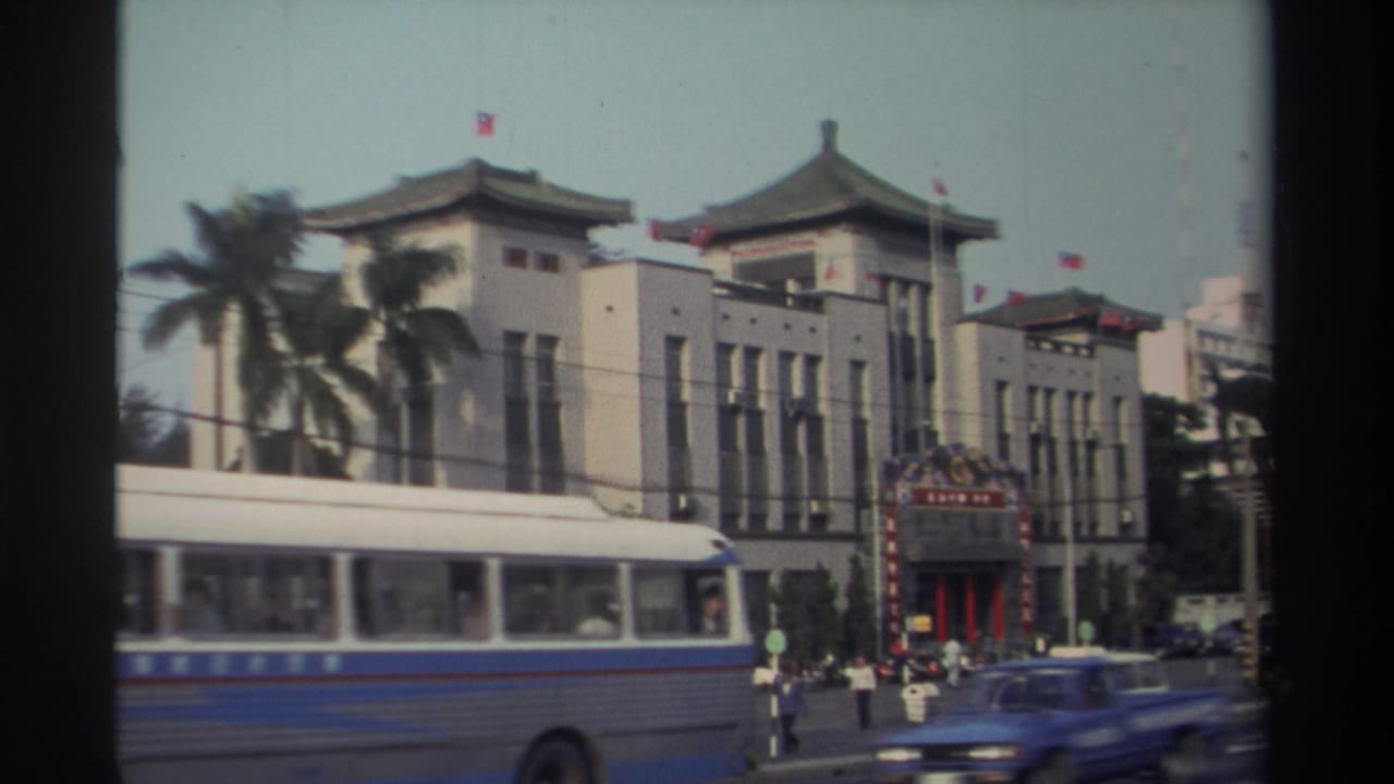 Bus and blue car pass in front of a flag-topped building on a city street.