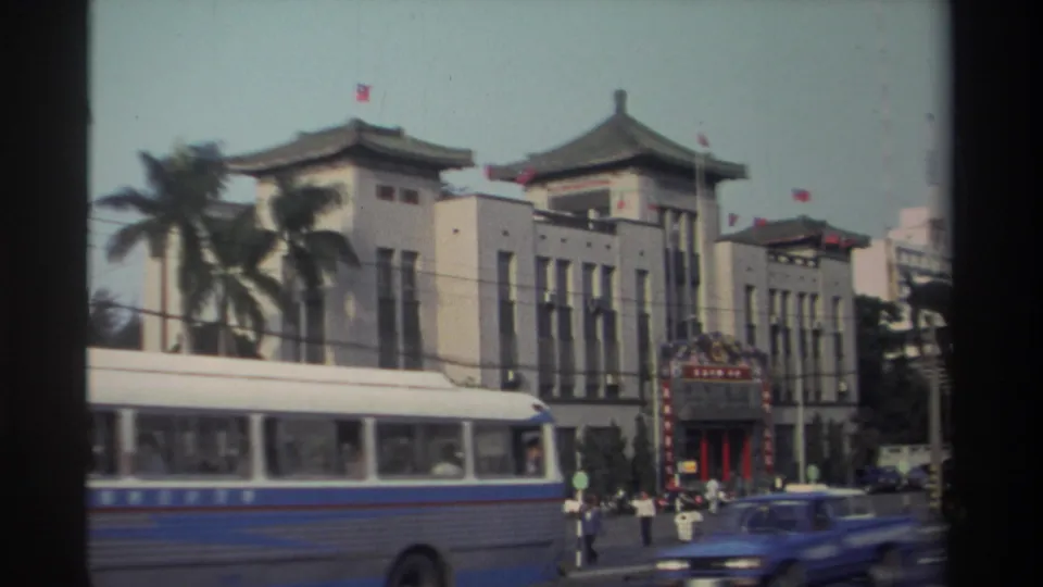 1982 Taiwan city traffic with a bus and blue car passing a traditional-style building with flags, palm trees, and pedestrians nearby.