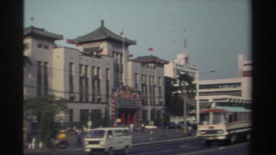 1982 Taiwan street scene with a white van, bus, scooters, and pedestrians beside a red entrance to a traditional-style building.