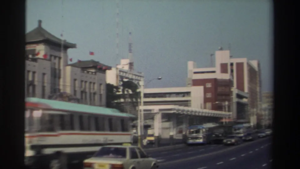 1982 Taiwan urban roadway with buses, cars, flags, and a traditional-style building set beside larger modern city structures.