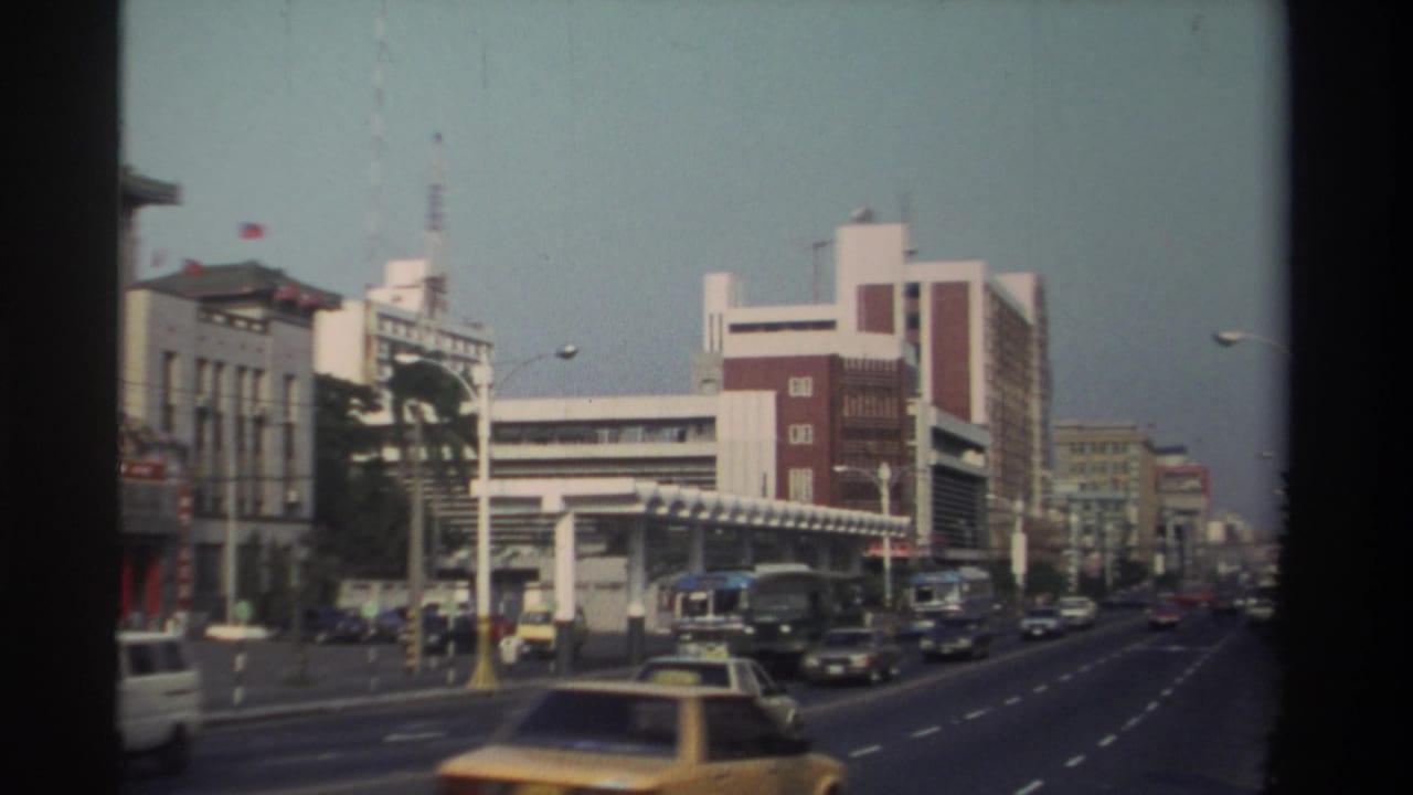 1982 Taiwan home-movie view of a busy city street with buses, cars, taxis, streetlights, and mid-rise commercial buildings.
