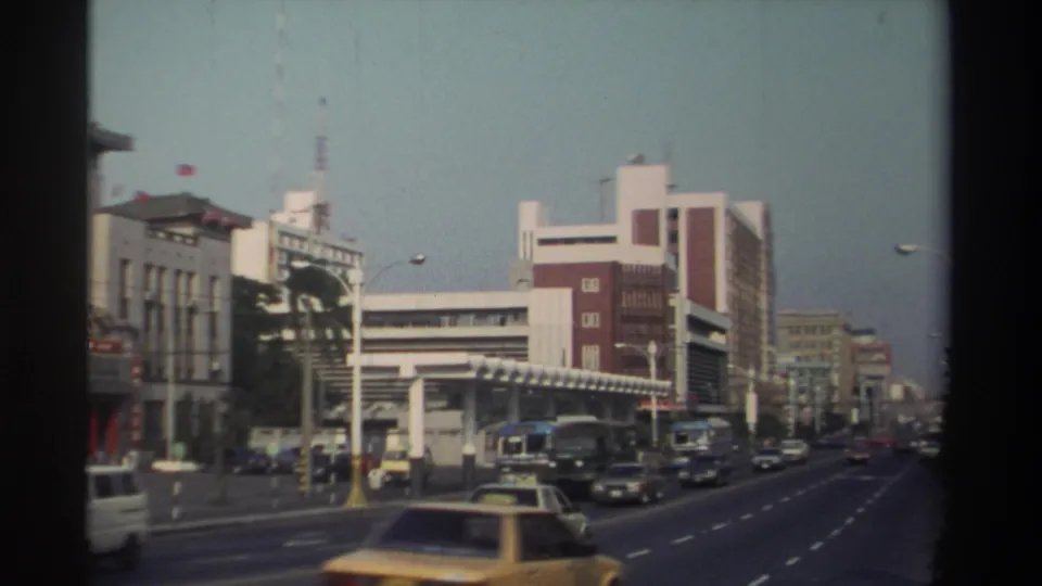 1982 Taiwan home-movie view of a busy city street with buses, cars, taxis, streetlights, and mid-rise commercial buildings.