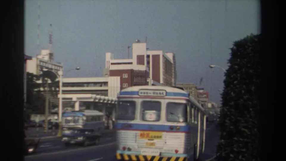 A bus seen from behind travels along a 1982 Taiwan city street, with another bus, cars, buildings, and dense roadside greenery visible.