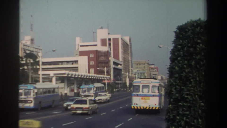 Traffic on a 1982 Taiwan city road includes buses, sedans, and taxis beside mid-rise buildings and a large roadside hedge.