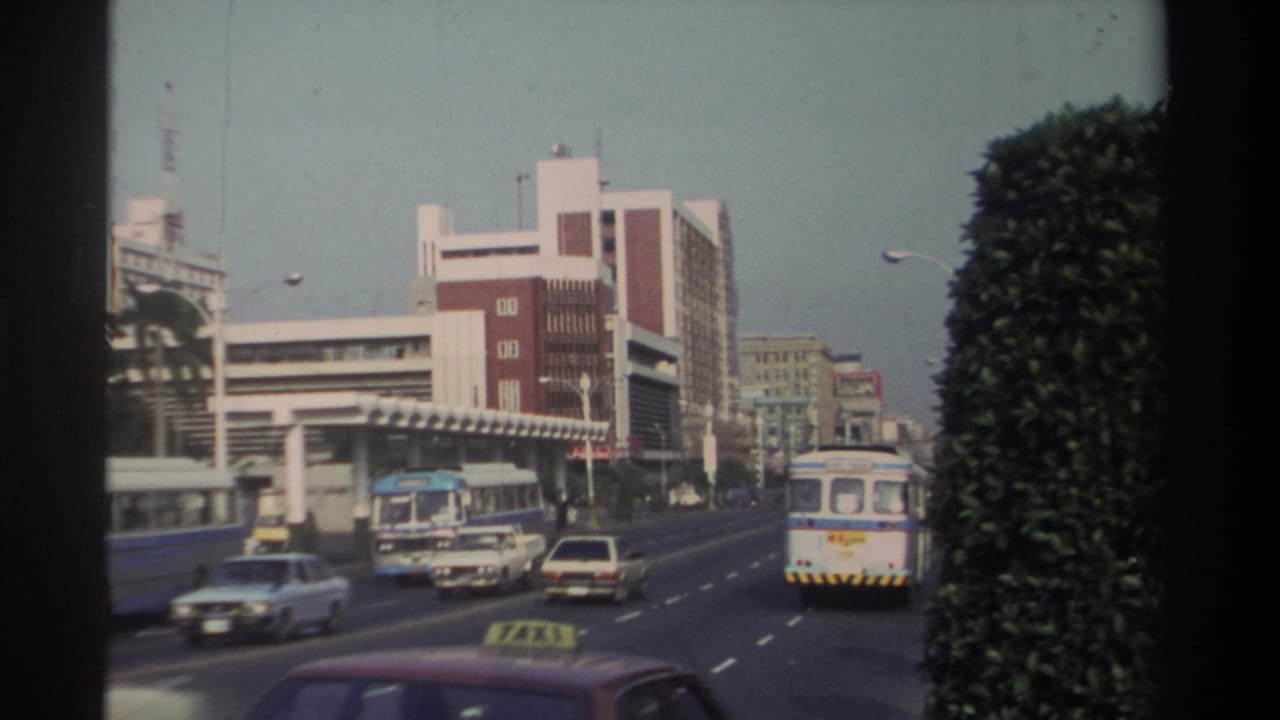 Multi-lane city road with buses, cars, a taxi roof sign, and a large hedge beside modern buildings.