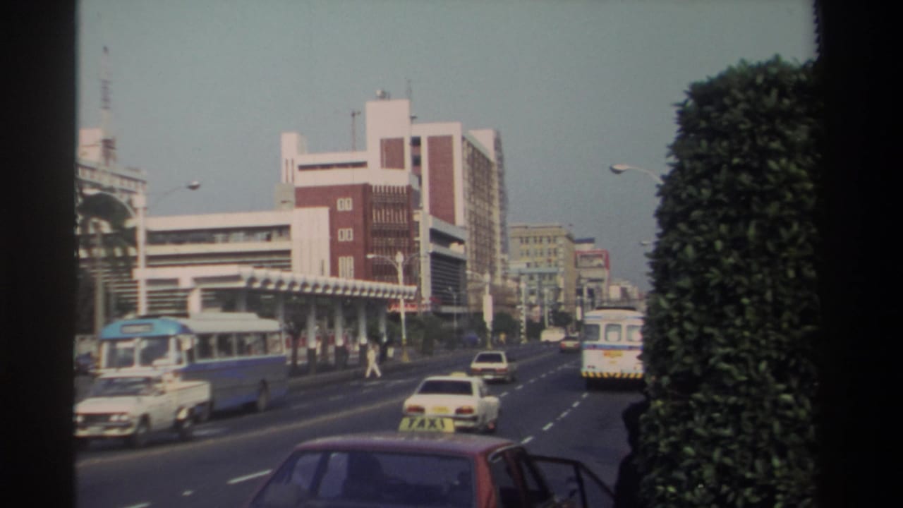 1982 Taiwan urban roadway scene with buses and cars moving past a prominent red-and-white building, roadside greenery, and a taxi in the foreground.