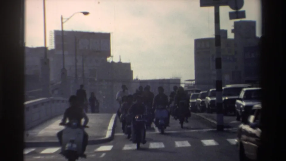 Street-level 1982 Taiwan view with scooter riders, pedestrians, parked cars, road markings, and buildings seen through soft exposure and motion blur.