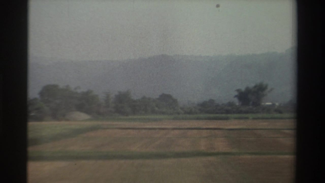 1982 Taiwan countryside view with agricultural fields, scattered trees, hazy hills, and dark window framing from a moving vehicle.