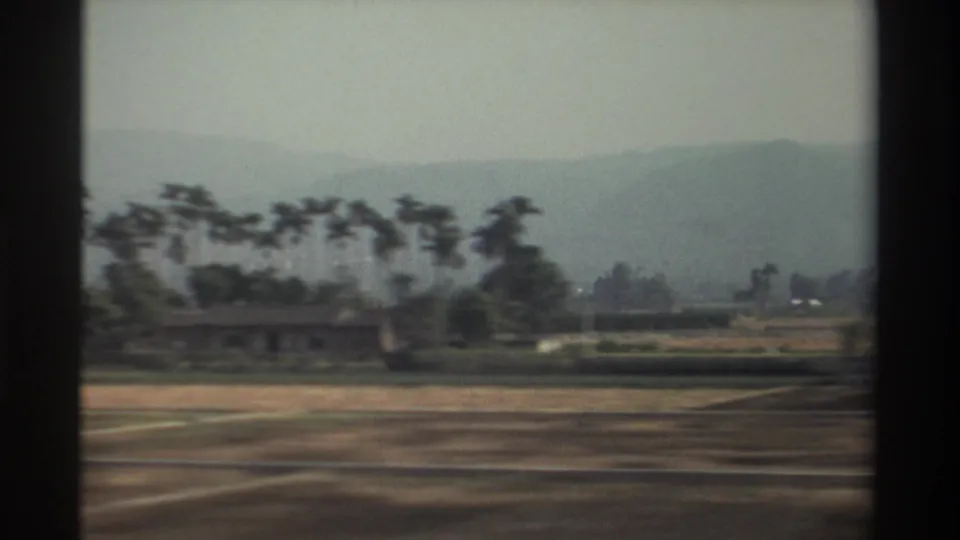 1982 Taiwan rural view with a low building among trees, cultivated fields, and a hazy mountain ridge under overcast light.