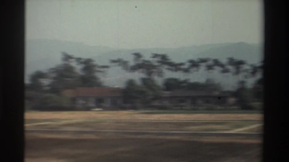 1982 Taiwan home-movie view from a moving vehicle, showing palm trees, low roadside buildings, open ground, and distant hazy hills.
