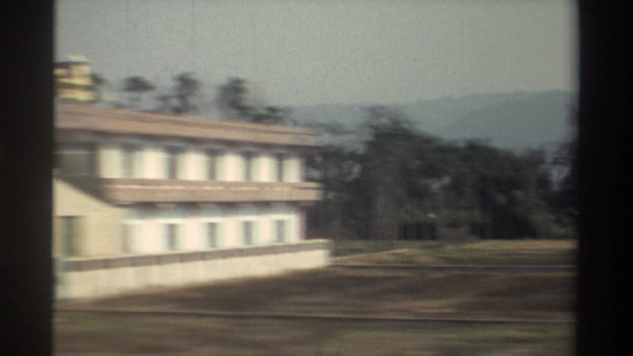White multi-story roadside building with trees and hills behind it.