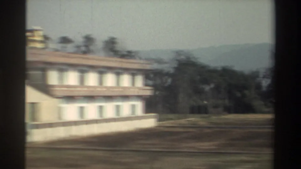 1982 Taiwan moving-vehicle frame with a white roadside building, trees, open verge, and hazy hills beyond.