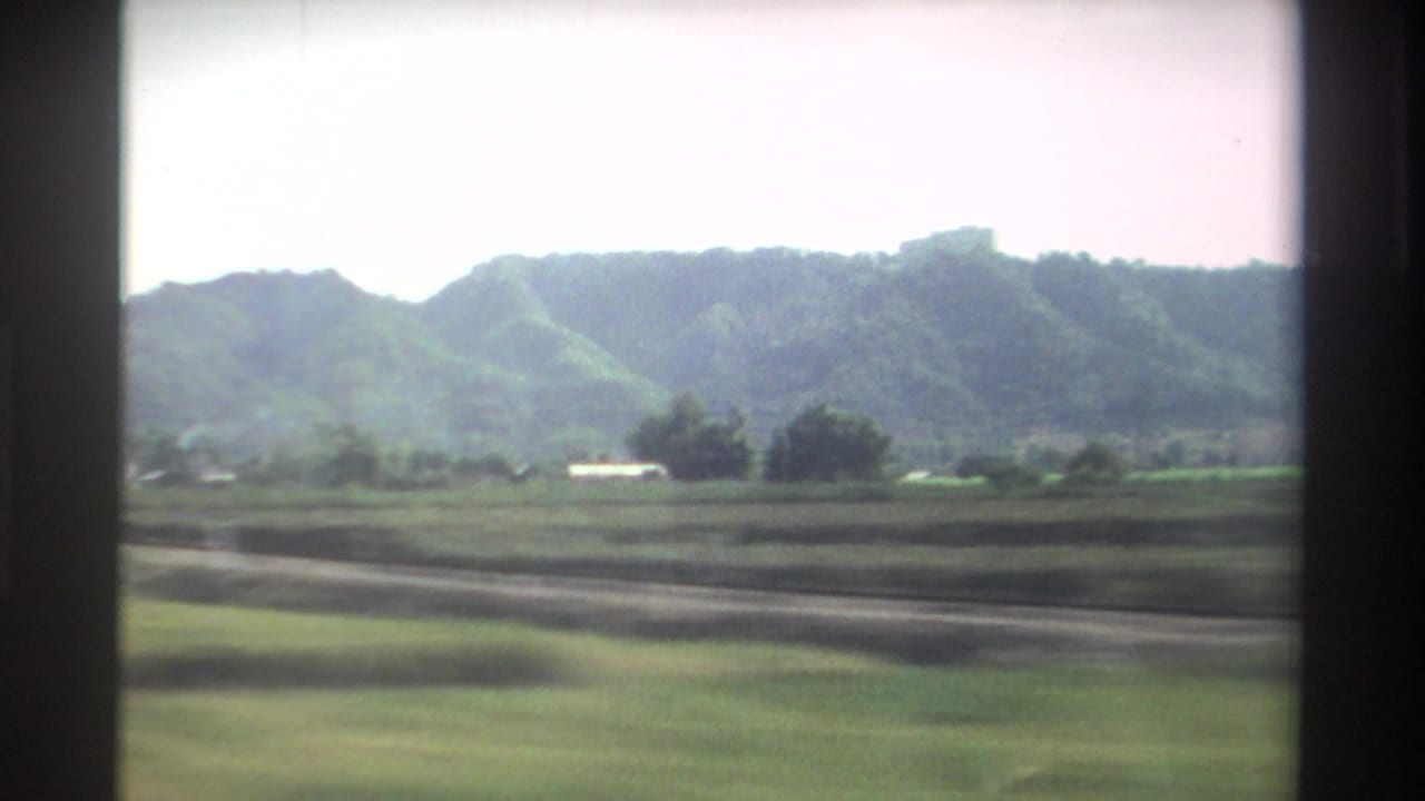Green fields and distant hills viewed through a vehicle window on a bright day.