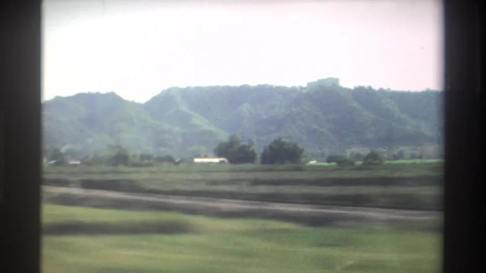 1982 Taiwan farmland passes by in bright daylight, with green fields, distant wooded hills, scattered trees, and visible window framing.