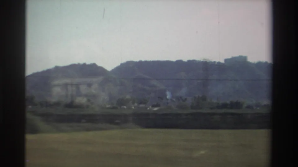 1982 Taiwan countryside seen from a car window, with open fields, low hills, scattered structures, and soft Super 8 color.