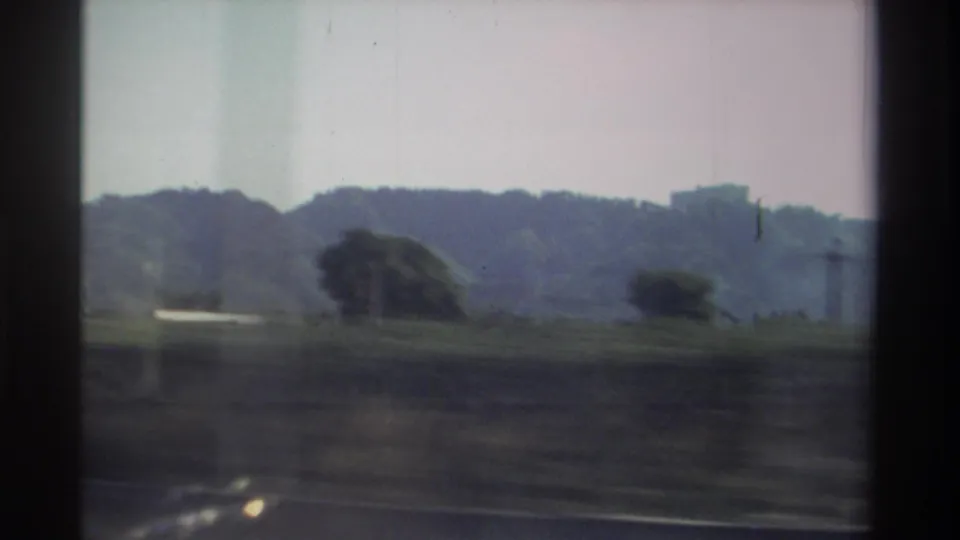 1982 Taiwan 8mm frame from a moving vehicle, showing roadside fields, trees, low hills, and a distant structure on the ridge.
