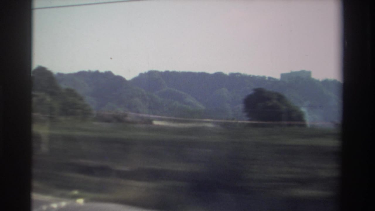1982 Taiwan home-movie frame showing green rural fields and wooded hills passing beyond a dark vehicle window edge.