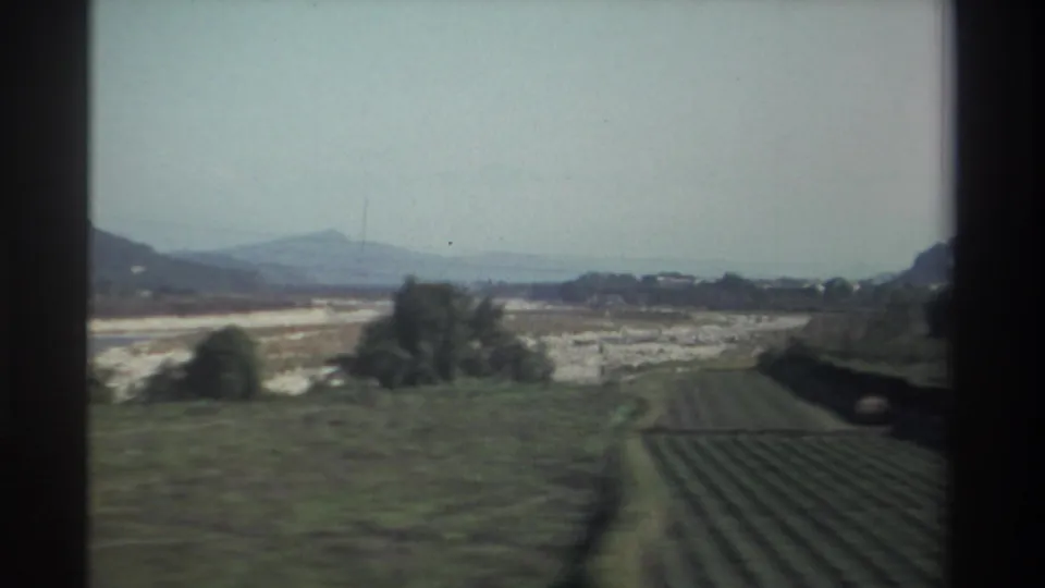 1982 Taiwan rural landscape with open fields, distant hills, roadside greenery, and a dark window edge from a moving vehicle.