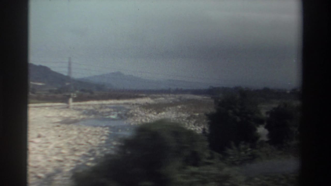 1982 Taiwan home-movie frame showing a rocky riverbed, distant hills, power lines, and dark vehicle-window edges.