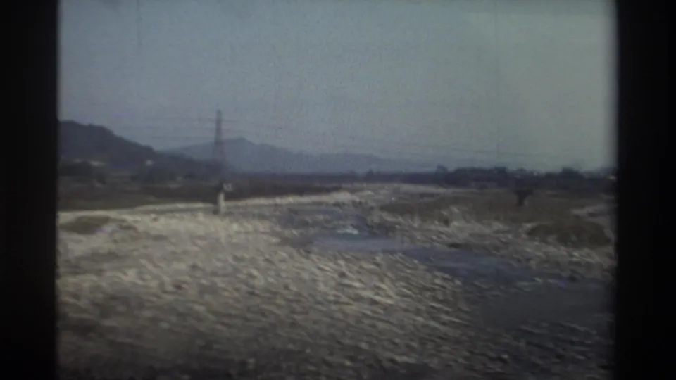 1982 Taiwan travel frame with a dry rocky riverbed, scattered vegetation, power lines, and hazy hills beyond.