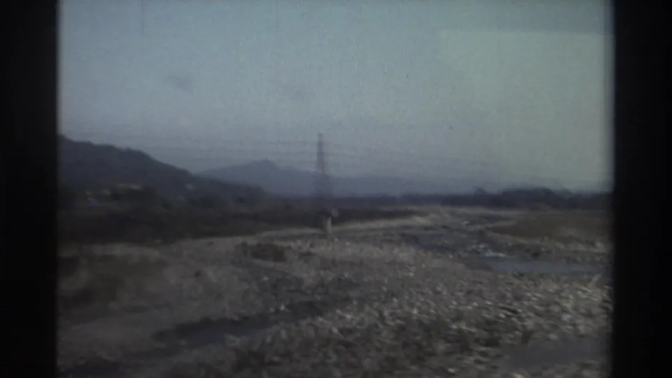 Soft 8mm frame from Taiwan in 1982 showing a rocky riverbed route, utility tower, hills, and motion-blurred foreground.