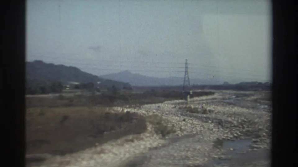 1982 Taiwan frame showing a rocky riverbed, shallow water, brush, a transmission tower, and dark window-edge framing.