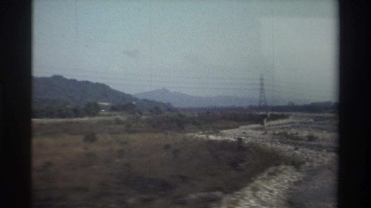 1982 Taiwan home-movie frame showing a dry riverbed across a rural valley with low hills, scrub vegetation, power lines, and a distant pylon.