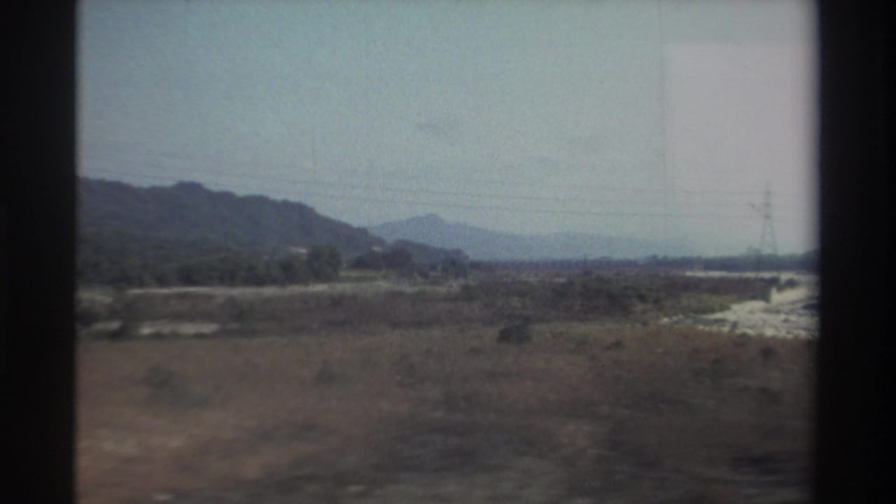 1982 Taiwan travel frame with open scrubland, hazy hills, a dry channel at right, and power infrastructure in the distance.