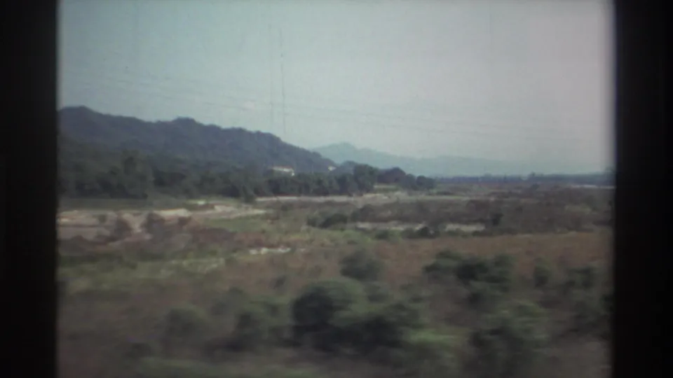1982 Taiwan frame showing shrubs in the foreground, a pale dry riverbed, low hills, and faint power lines across the sky.