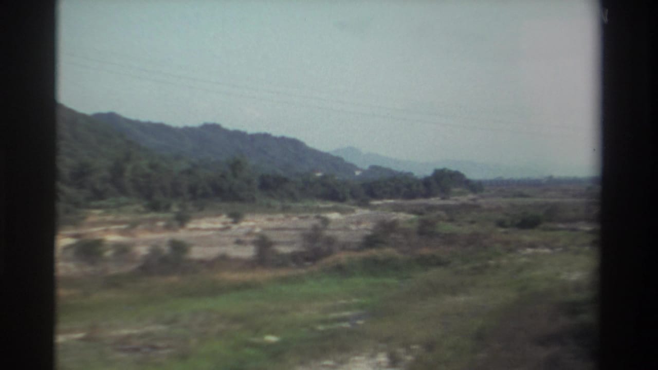 Riverbed or open ground with wooded hills beyond the train window.