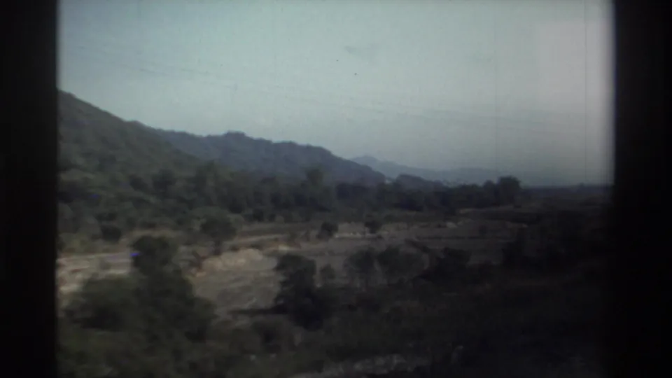 Dim 1982 Taiwan view from a train window, with hills, fields, trees, and distant ridgelines softened by motion.
